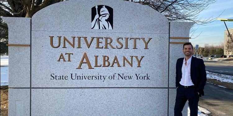 Entrevista com o Aluno Patricio Jousset, da SUNY O aluno Patricio está, de pé, sorrindo ao lado de uma parede de concreto que contém o logo da SUNY Albany