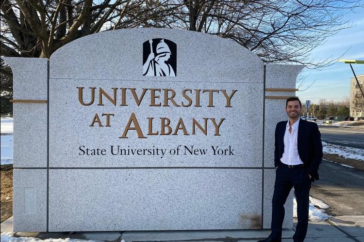 O aluno Patricio está, de pé, sorrindo ao lado de uma parede de concreto que contém o logo da SUNY Albany